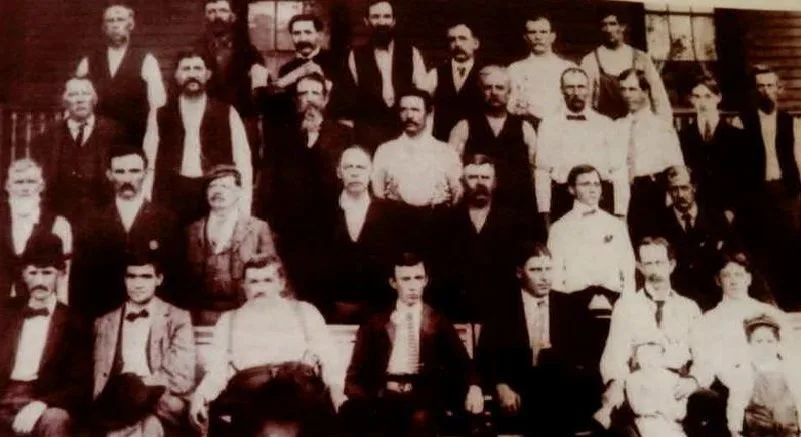 A large group of men in vests and ties posed on the steps of a porch