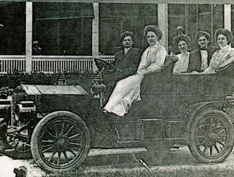 Five women seated in an early open automobile in front of a resort porch